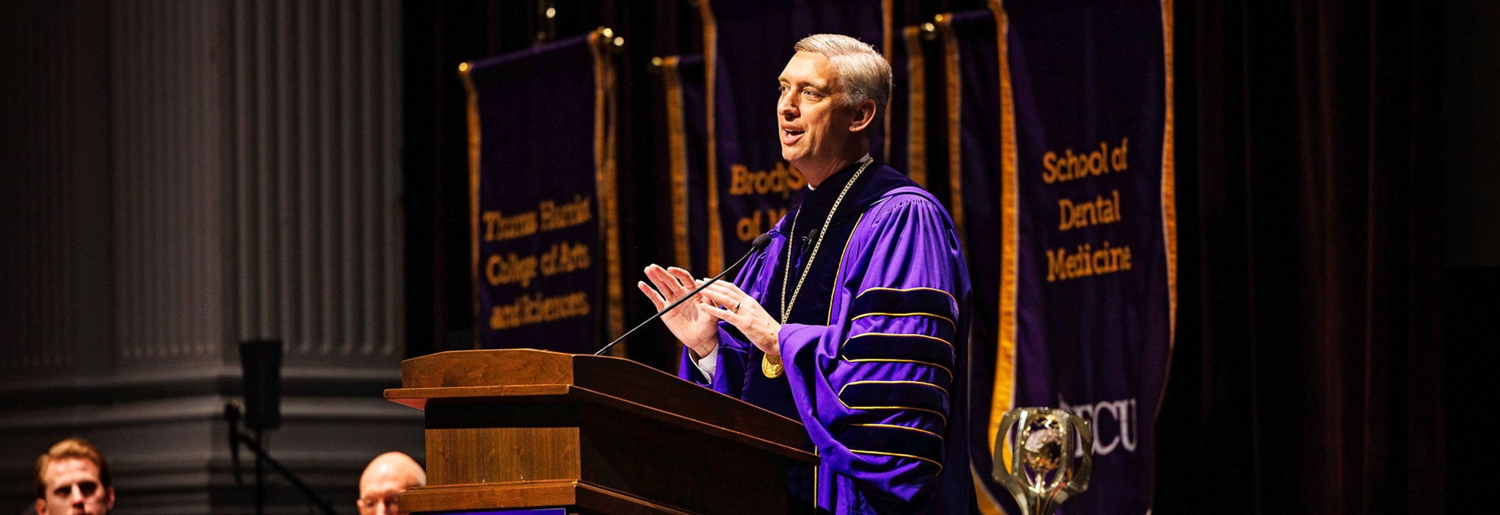 ECU Chancellor Philip Rogers speaks at a podium during a formal academic event, wearing purple regalia, with banners for various ECU colleges in the background.